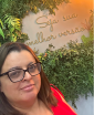 Mulher de cabelos lisos castanhos e óculos de grau posando em frente a uma parede decorada com folhagens e a frase 'Seja sua melhor versão' em letras douradas