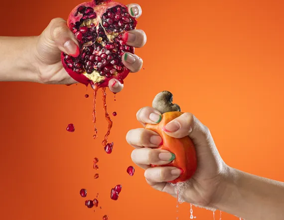 Duas mãos espremem frutas em frente a um fundo laranja: à esquerda, uma romã aberta com sementes e suco escorrendo; à direita, um caju sendo pressionado, liberando seu líquido.