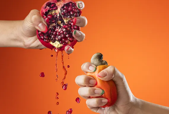 Duas mãos espremem frutas em frente a um fundo laranja: à esquerda, uma romã aberta com sementes e suco escorrendo; à direita, um caju sendo pressionado, liberando seu líquido.