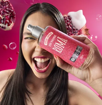 A imagem mostra uma mulher sorrindo e segurando um frasco de perfume com o rótulo "ROMÃ". Ao fundo, há sementes de romã e um fundo colorido em tons de rosa.