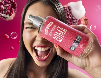 A imagem mostra uma mulher sorrindo e segurando um frasco de perfume com o rótulo "ROMÃ". Ao fundo, há sementes de romã e um fundo colorido em tons de rosa.