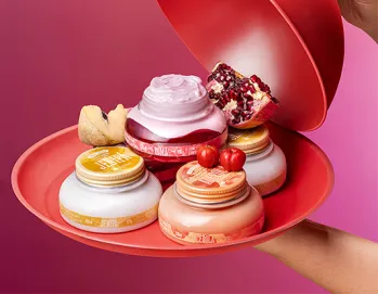 A imagem mostra uma mão segurando uma bandeja vermelha com diversos potes de cremes, incluindo fragrâncias de romã, cereja e outras, enquanto uma tampa vermelha está sendo levantada. Ao fundo, o cenário é colorido em tons de rosa.