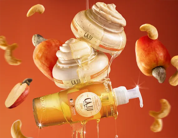 Dois potes de creme corporal e um frasco de óleo com rótulo de caju empilhados, cobertos por um fio de óleo dourado escorrendo, sobre fundo laranja vibrante. A composição destaca brilho e textura dos produtos, transmitindo sensação de nutrição e cuidado.