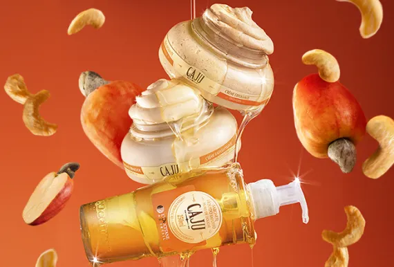 Dois potes de creme corporal e um frasco de óleo com rótulo de caju empilhados, cobertos por um fio de óleo dourado escorrendo, sobre fundo laranja vibrante. A composição destaca brilho e textura dos produtos, transmitindo sensação de nutrição e cuidado.
