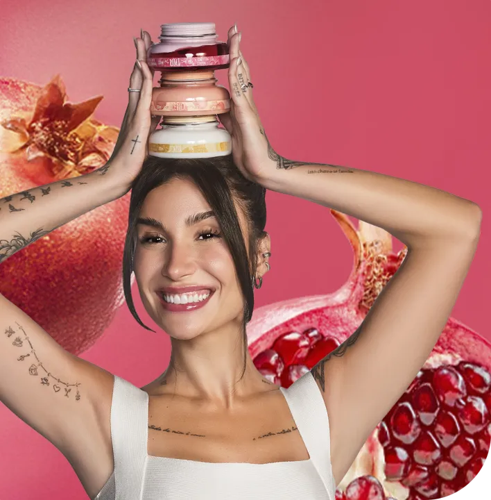 A imagem mostra uma mulher sorrindo e segurando uma pilha de potes de cremes sobre a cabeça. Ela tem cabelo escuro e curto, com tatuagens visíveis nos braços, e está em um fundo rosa com romãs ao fundo