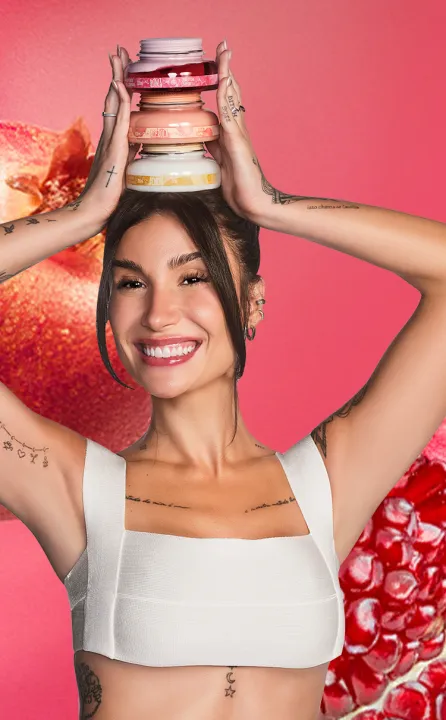 A imagem mostra uma mulher sorrindo e segurando uma pilha de potes de cremes sobre a cabeça. Ela tem cabelo escuro e curto, com tatuagens visíveis nos braços, e está em um fundo rosa com romãs ao fundo