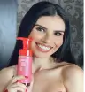 Mulher de cabelos longos e pretos, com maquiagem e unhas vermelhas, sorrindo enquanto segura um frasco de mousse de banho com rótulo rosa e vermelho, em ambiente interno com parede decorada ao fundo.