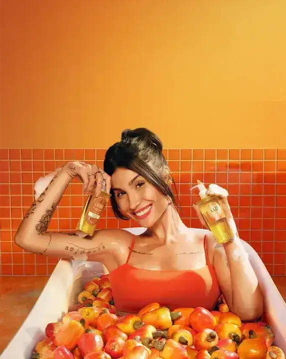 Mulher sorridente em uma banheira cheia de cajus, vestindo roupa laranja, segurando dois frascos de produtos cosméticos nas mãos, com parede de azulejos laranja ao fundo e ambiente em tons quentes