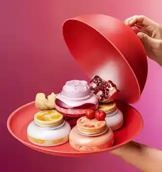 A imagem mostra uma mão segurando uma bandeja vermelha com diversos potes de cremes, incluindo fragrâncias de romã, cereja e outras, enquanto uma tampa vermelha está sendo levantada. Ao fundo, o cenário é colorido em tons de rosa.