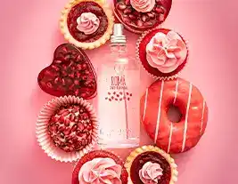 A imagem mostra um frasco de perfume no centro, cercado por doces decorados em tons de rosa e vermelho sobre um fundo rosa. Entre os doces, há cupcakes, tortinhas, um donut e um bombom em forma de coração.