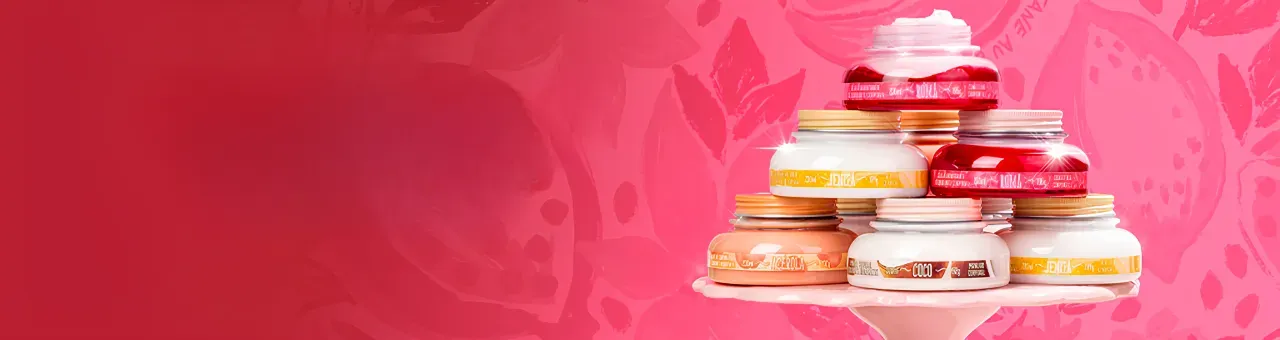 A imagem mostra uma pilha de potes de cremes em uma base branca, com diferentes fragrâncias, como "ROMÃ", "COCO" e "JENIPAPO", sobre um fundo rosa vibrante com desenhos de romã. Ao lado, há um texto em branco e rosa, escrito "Vem EMPREENDER COMIGO, mon amour!".