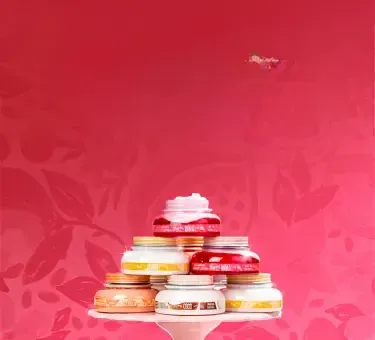 A imagem mostra uma pilha de potes de cremes em uma base branca, com diferentes fragrâncias, como "ROMÃ", "COCO" e "JENIPAPO", sobre um fundo rosa vibrante com desenhos de romã. Ao lado, há um texto em branco e rosa, escrito "Vem EMPREENDER COMIGO, mon amour!".